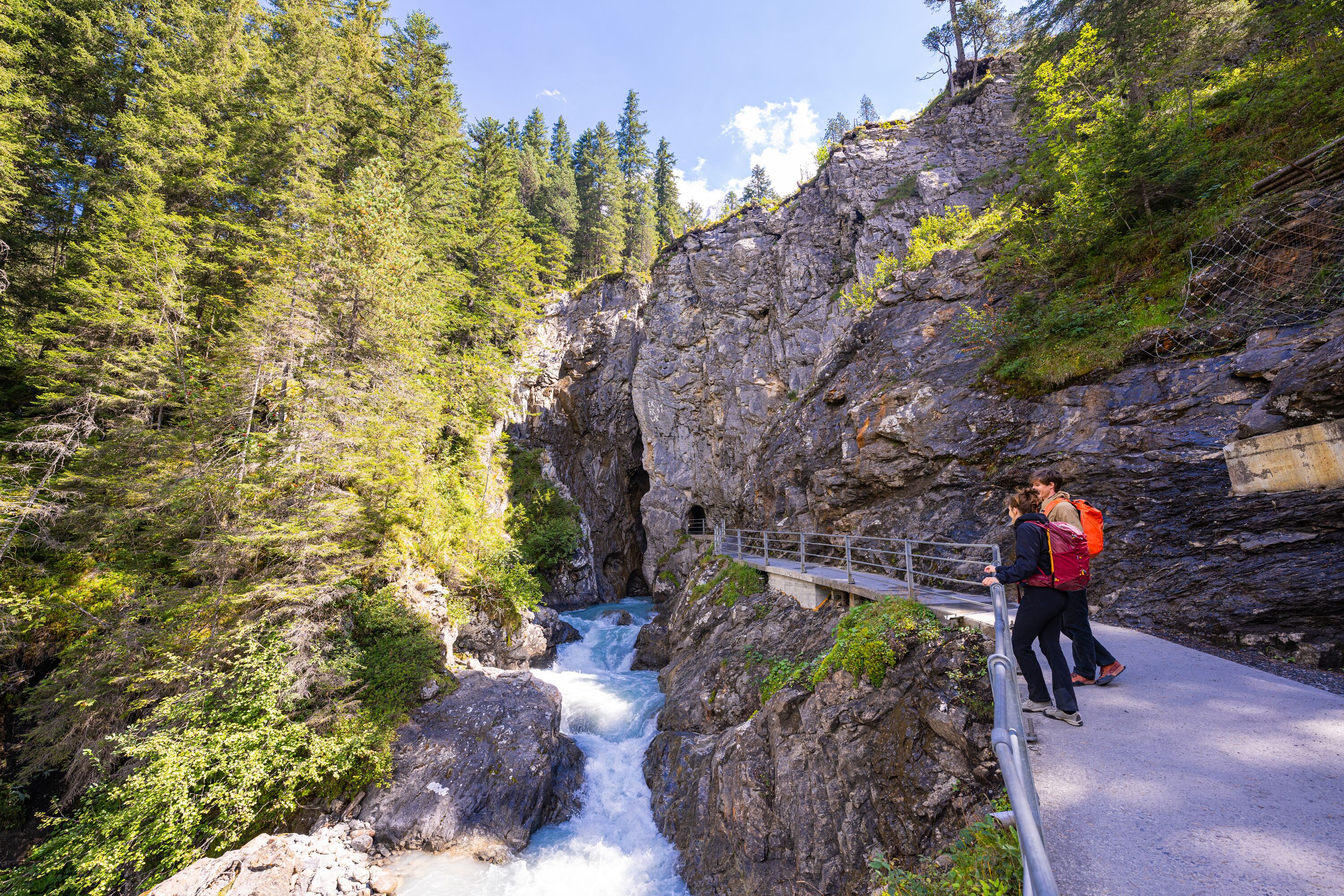 Gletscherschlucht Rosenlaui Der Fluss formte die Rosenlaui Gletscherschlucht über Jahrhunderte. Der Weg führt an der Felswand über dem Fluss in die Schlucht hinein. Zwei Wanderer bestaunen den Fluss vor dem Eingang in die Schlucht.